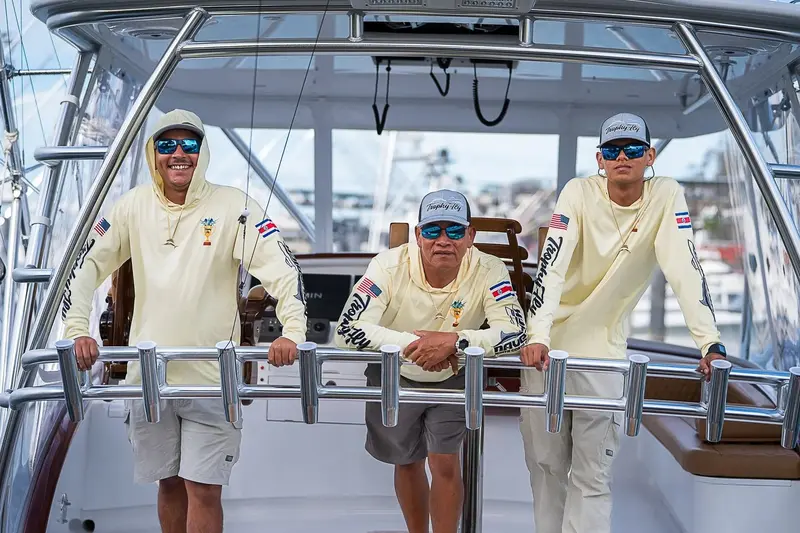 Tres tripulantes con camisas de manga larga y gorras, de pie en la cabina de un barco de pesca
