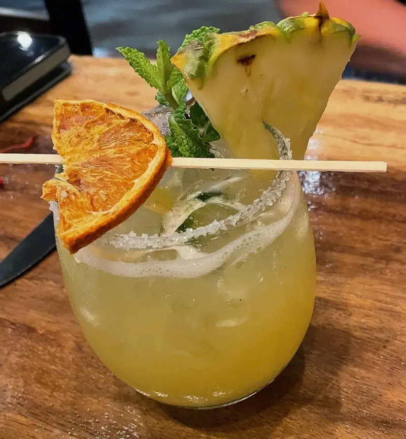 Cóctel amarillo con el borde escarchado, rodaja de naranja deshidratada, piña y hierbabuena sobre una mesa de madera