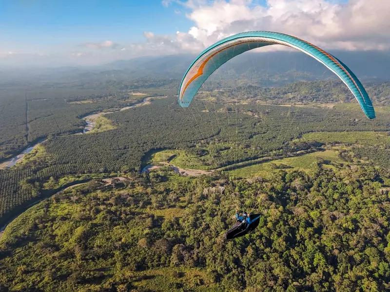 Vista aérea de un valle con un río entre bosque y un parapente turquesa en vuelo