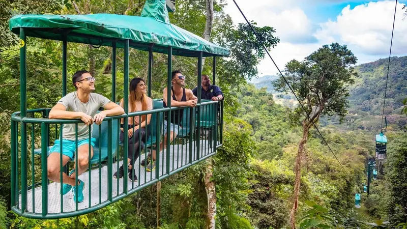 Cabina verde del teleférico con visitantes sobre el bosque, con colinas al fondo