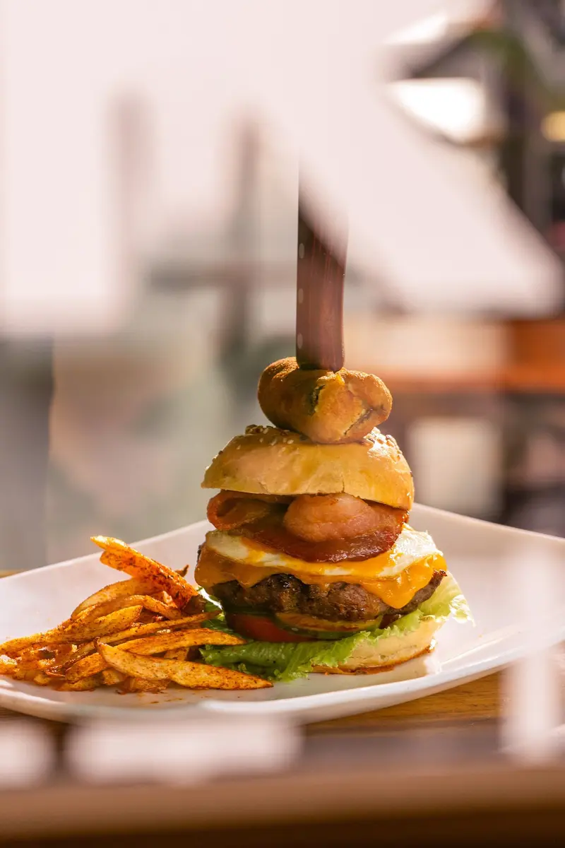Hamburguesa alta con queso derretido y tomate, coronada con un pincho y servida con papas fritas