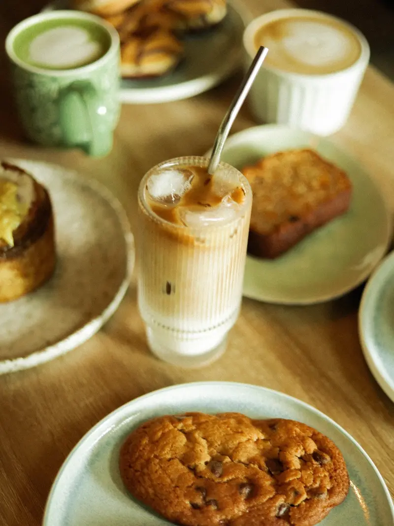 Latte frío en vaso alto, capuchino, pan de banano y una galleta sobre la mesa