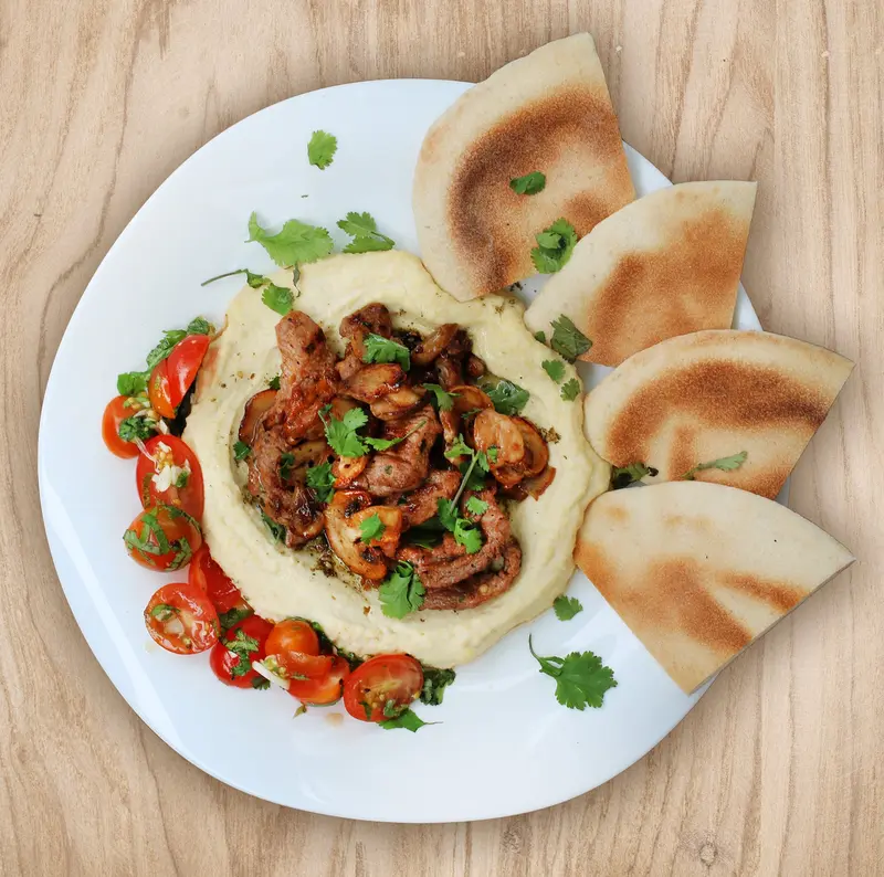 Hummus con carne y hongos salteados sobre un plato blanco, con pan de pita y tomates cherry
