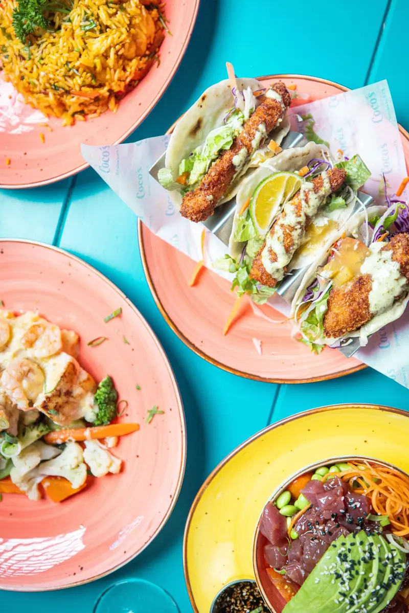 Mesa vista desde arriba con tacos de pescado, platos de pollo con verduras y un bowl de poke sobre una mesa celeste