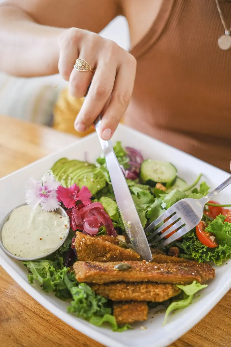 Mano con cuchillo y tenedor sobre una ensalada con aguacate, pepino, tomate cherry y filetes empanizados