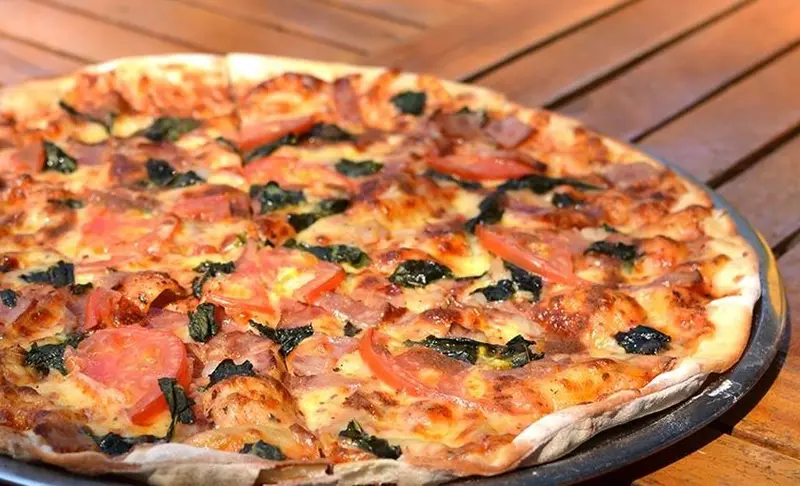 Pizza de masa fina con jamón, tomate y espinaca sobre una bandeja, en una mesa de madera