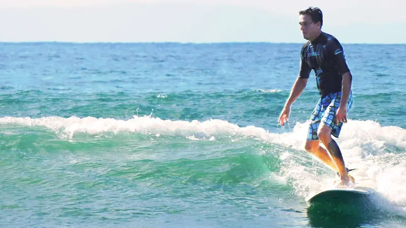 Surfista de pie sobre una tabla verde en una ola rompiendo, con el mar turquesa detrás