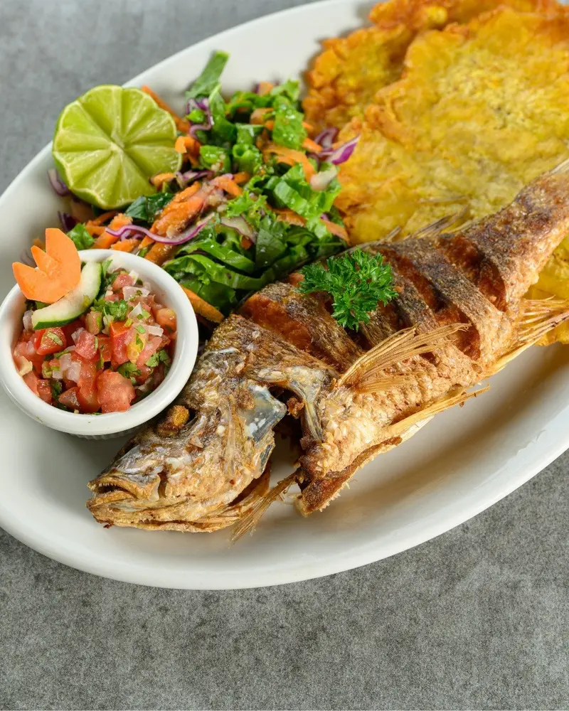 Pargo entero frito con patacones, ensalada, pico de gallo y una rodaja de limón en un plato ovalado