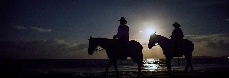 Dos jinetes a caballo en silueta contra la luna llena sobre el mar