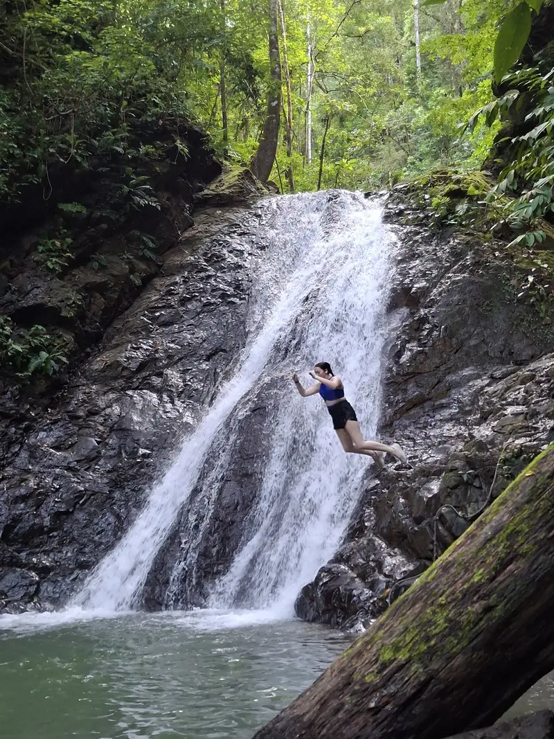 Excursionista saltando junto a una cascada de roca oscura, rodeada de selva y con una poza abajo