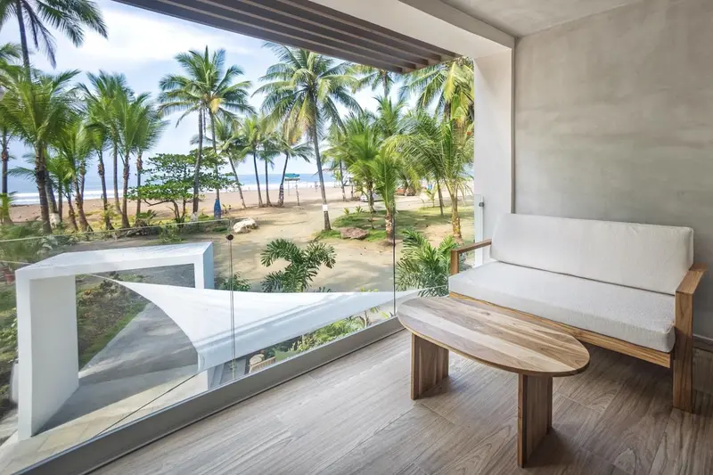 Balcón de una habitación con sillón blanco y mesa baja de madera, con vista a la playa y las palmeras