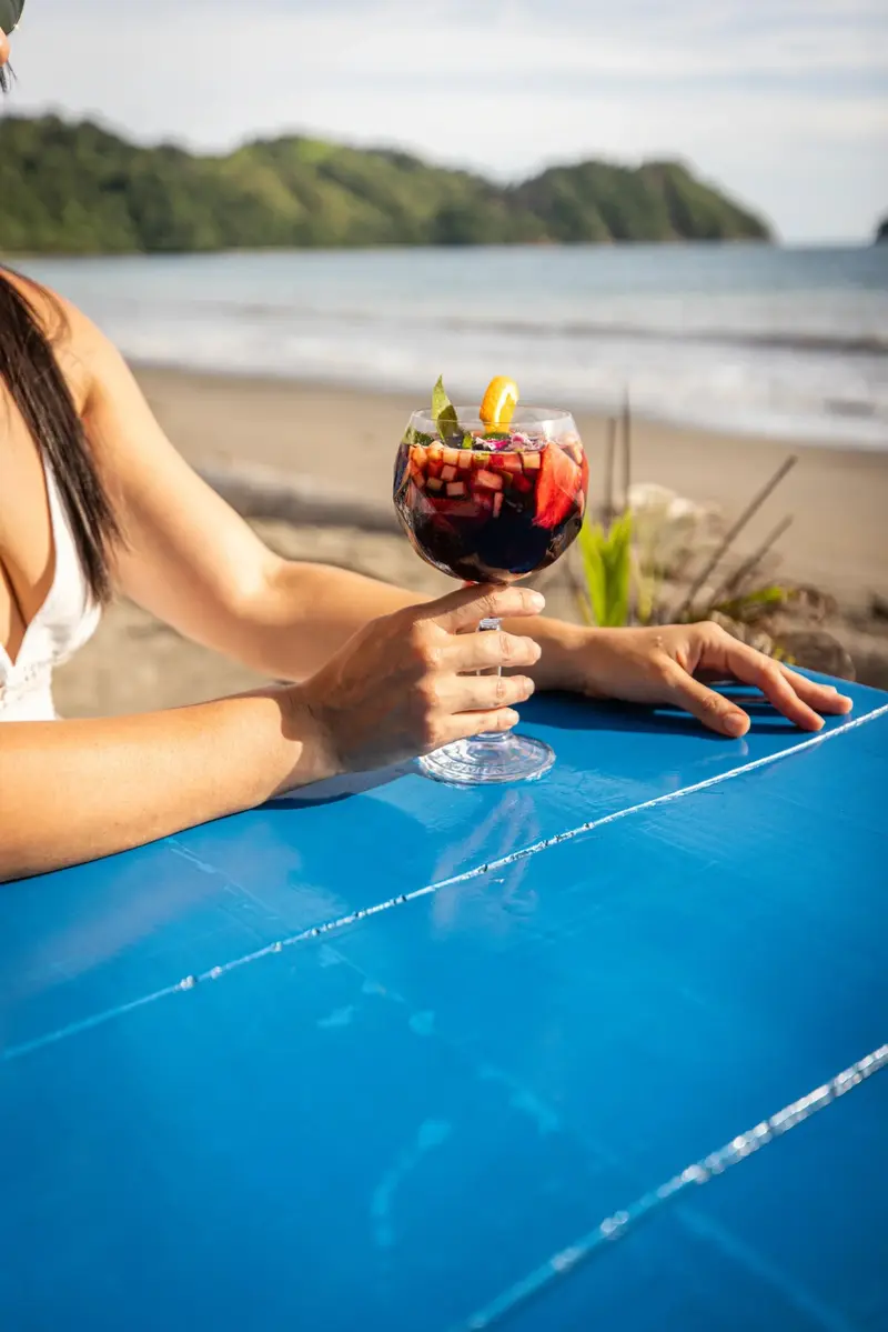 Mano sosteniendo una copa de sangría sobre una mesa azul, con una bahía y una isla al fondo