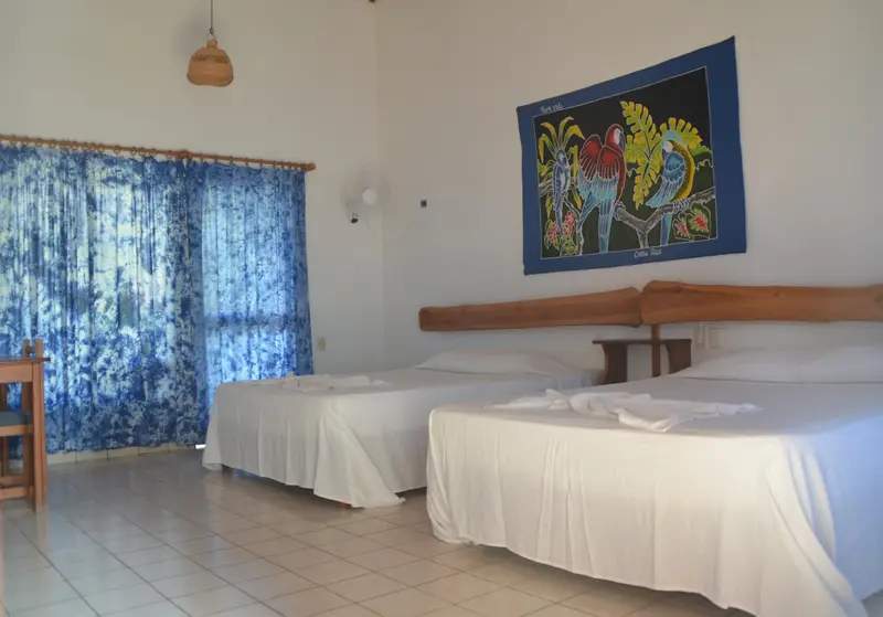 Habitación con dos camas dobles de sábanas blancas, cabecera de madera y un tapiz de colores en la pared