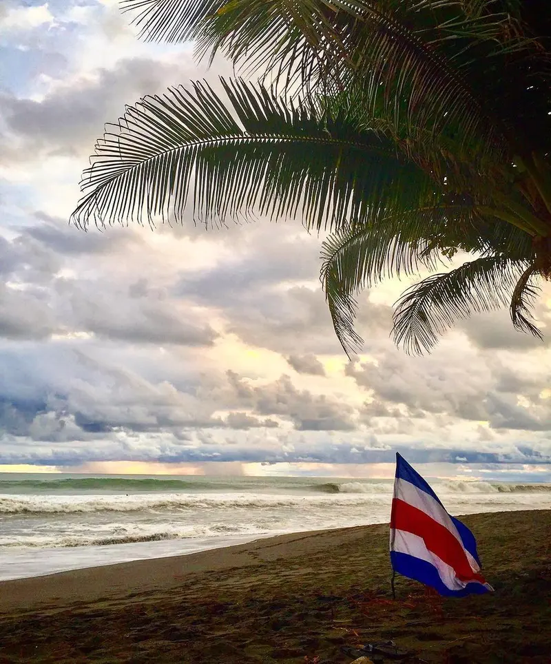 Playa de arena oscura con una bandera de Costa Rica y una palmera bajo un cielo de nubes