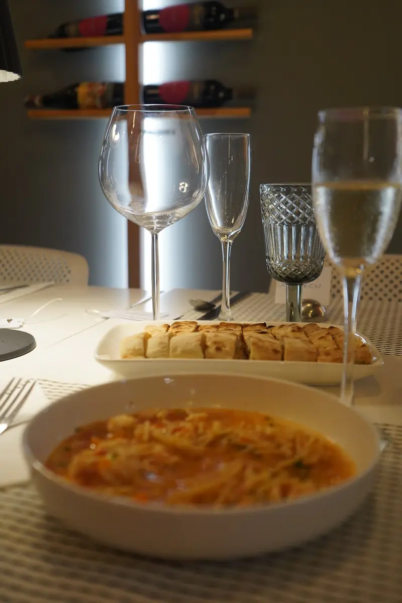Mesa con un plato hondo de sopa de pasta, focaccia cortada y copas, frente a una estantería de vinos