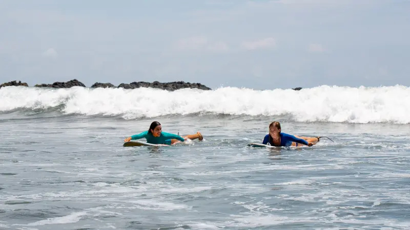 Dos personas acostadas sobre tablas en el agua, remando entre olas pequeñas