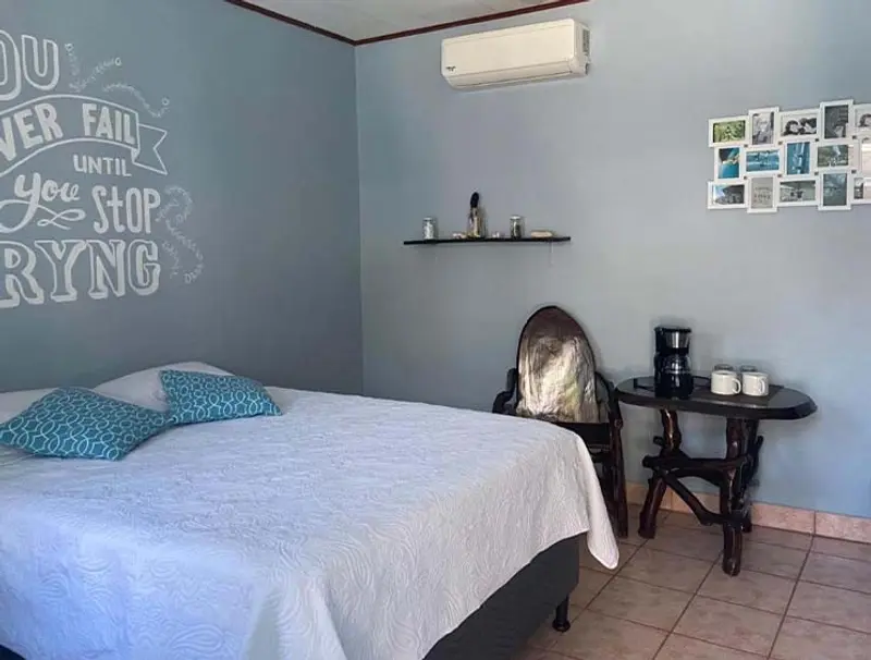 Habitación con cama doble de ropa blanca, aire acondicionado, mesa redonda con cafetera y pared azul con frase escrita