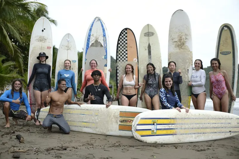 Grupo de alumnos con sus tablas de surf sobre la arena, frente a una hilera de tablas de pie