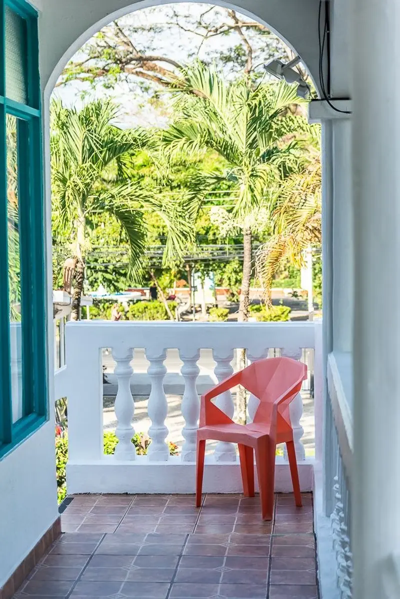 Balcón con silla roja y puerta turquesa que da a un jardín de palmeras