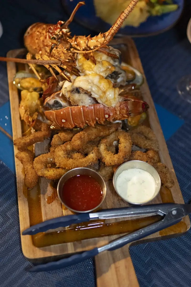 Mariscada servida en una tabla de madera con langosta, aros de calamar y salsas, sobre un mantel azul