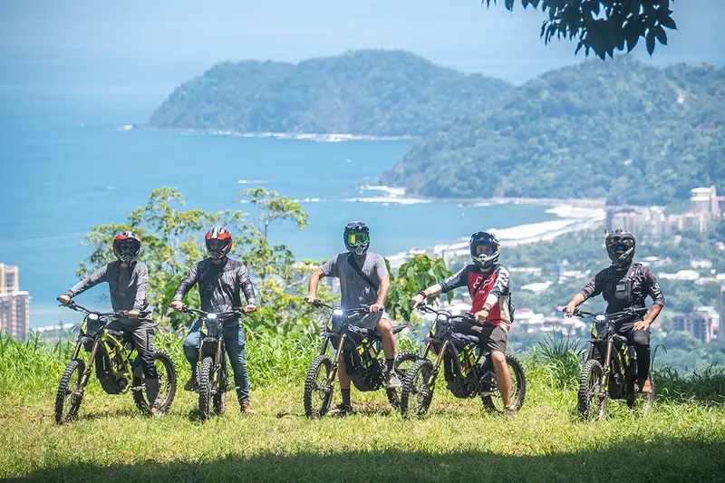 Cinco pilotos con casco junto a sus motos en un mirador con la bahía de Jacó al fondo