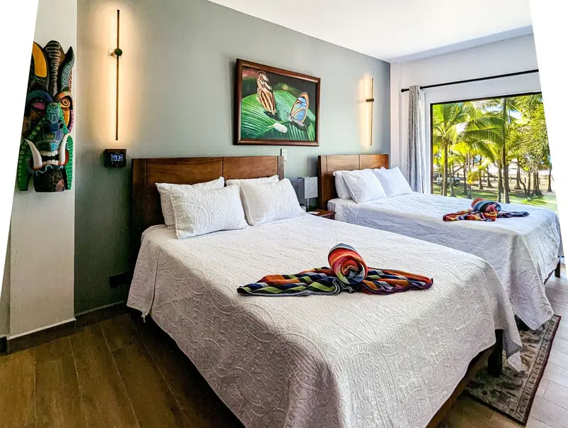 Habitación con dos camas queen y puerta de vidrio al balcón con vista a las palmeras