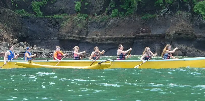 Canoa outrigger amarilla con remeros junto a un acantilado de roca y selva