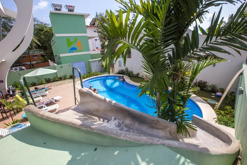 Piscina con tobogán de agua entre palmeras y jardín, frente al edificio del hostel