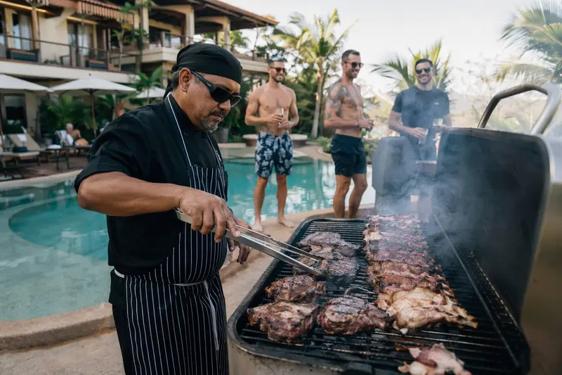 Chef con delantal asando carne en una parrilla junto a la piscina de una villa