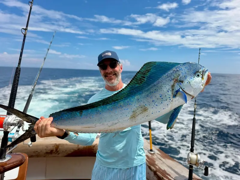 Pescador sonriente sosteniendo un dorado grande a bordo, con cañas a los lados y el mar al fondo
