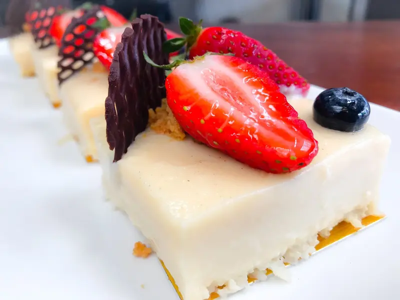 Postre cuadrado de crema blanca sobre base de galleta, con fresa, arándano y láminas de chocolate