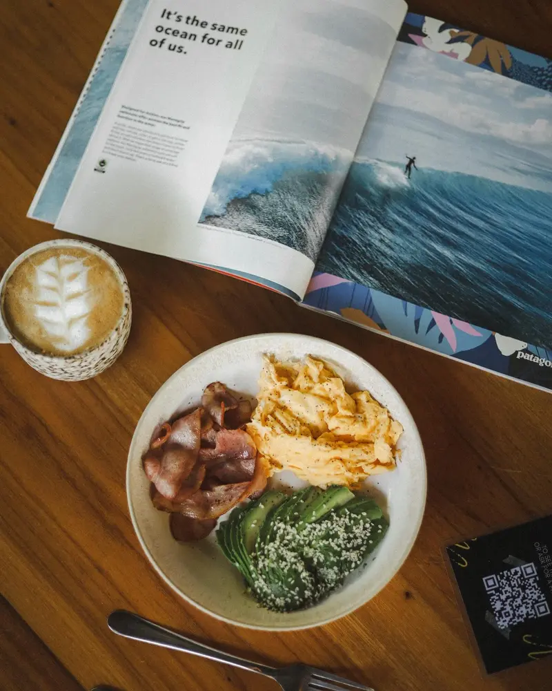 Plato de desayuno y capuchino con arte latte junto a una revista de surf abierta