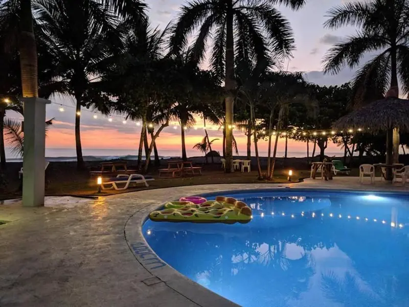 Piscina al atardecer con palmeras, luces encendidas y el mar al fondo