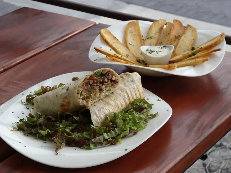 Burrito cortado por la mitad sobre hojas verdes, con una canasta de papas en bastón y aderezo