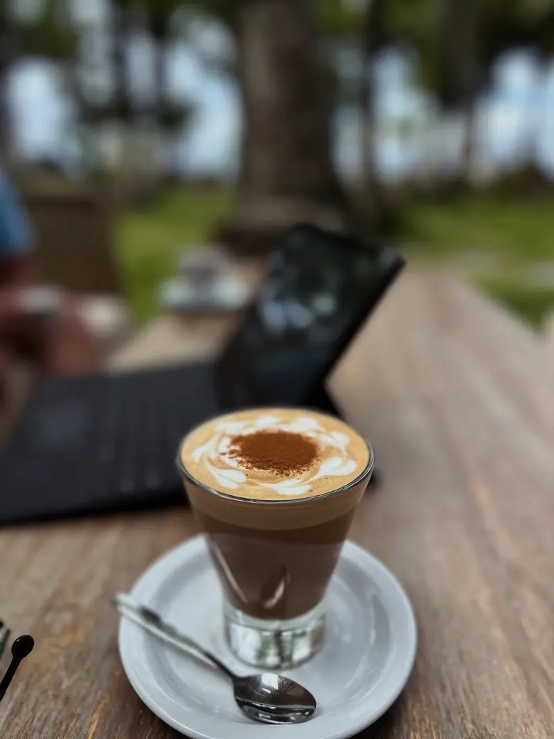 Café en vaso de vidrio sobre un plato con cuchara, con una laptop y palmeras desenfocadas al fondo