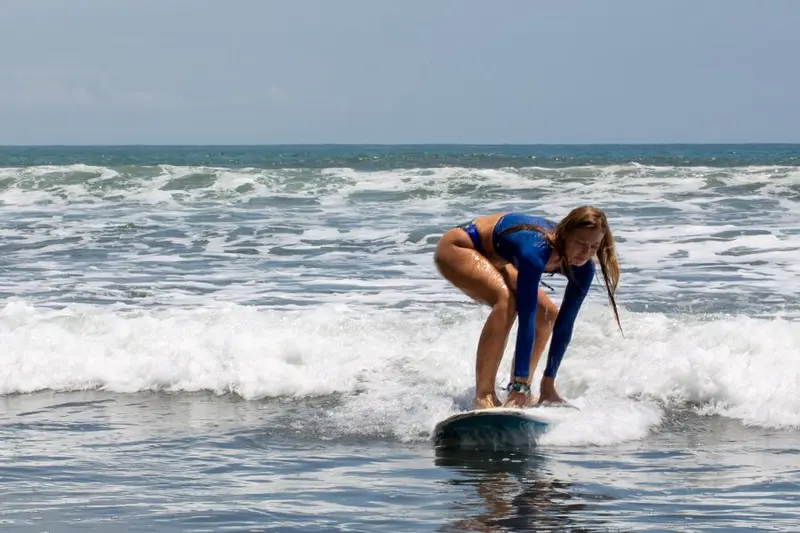 Persona agachada sobre una tabla de surf en el agua, con olas pequeñas al fondo