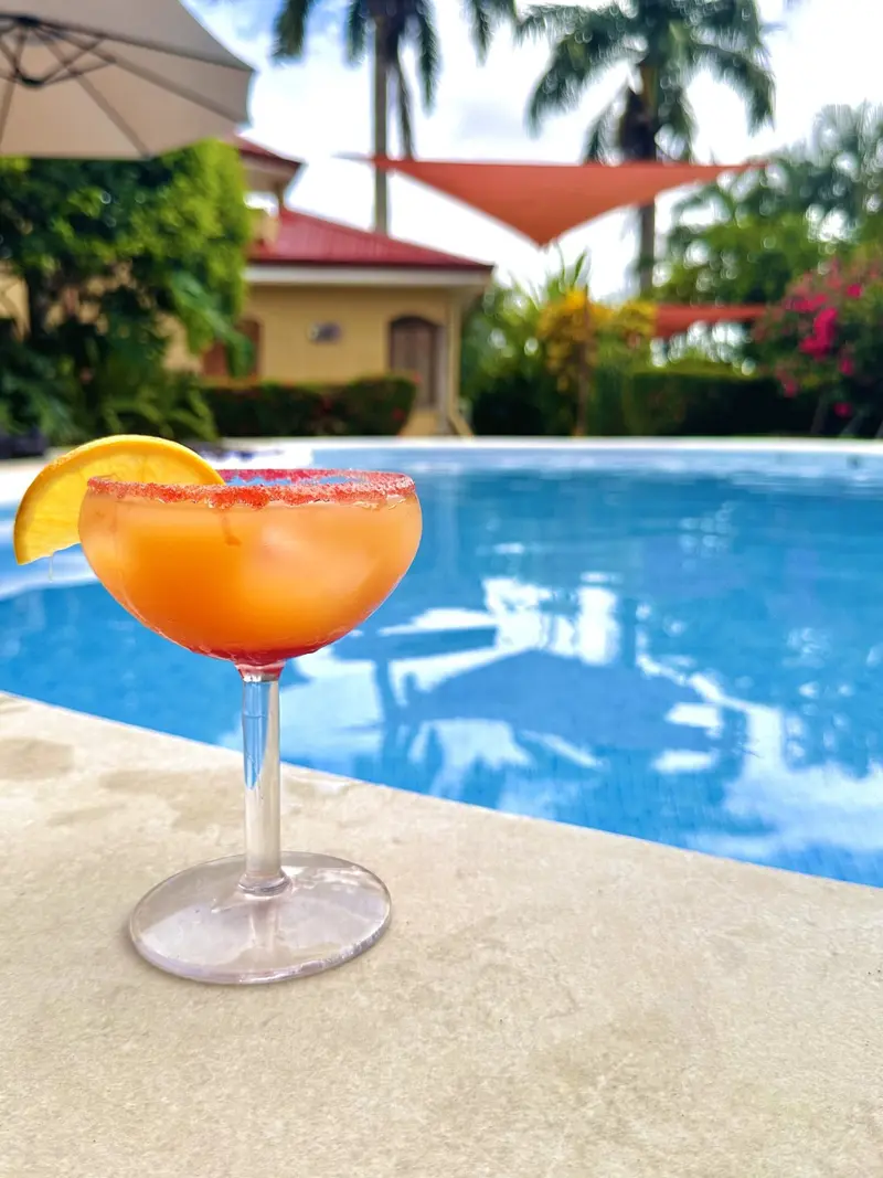 Cóctel naranja en una copa sobre el borde de la piscina, con jardín y villas al fondo