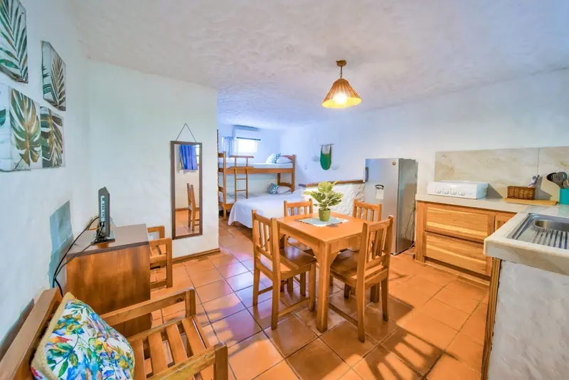 Sala-comedor de un estudio con mesa de madera, sofá con cojines turquesa y cocina al fondo