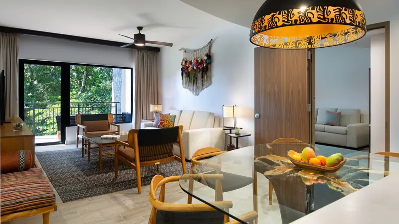Sala de una villa con sofás, mesa de comedor de vidrio, lámpara de mimbre y puertas corredizas hacia el balcón