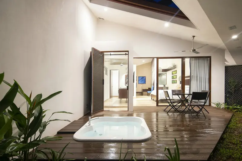 Jacuzzi sobre una terraza de madera junto a una mesa con sillas, con la habitación y la ducha al fondo