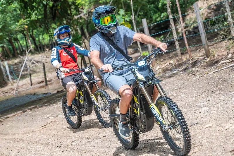 Dos pilotos con casco en motos eléctricas sobre un camino con vegetación