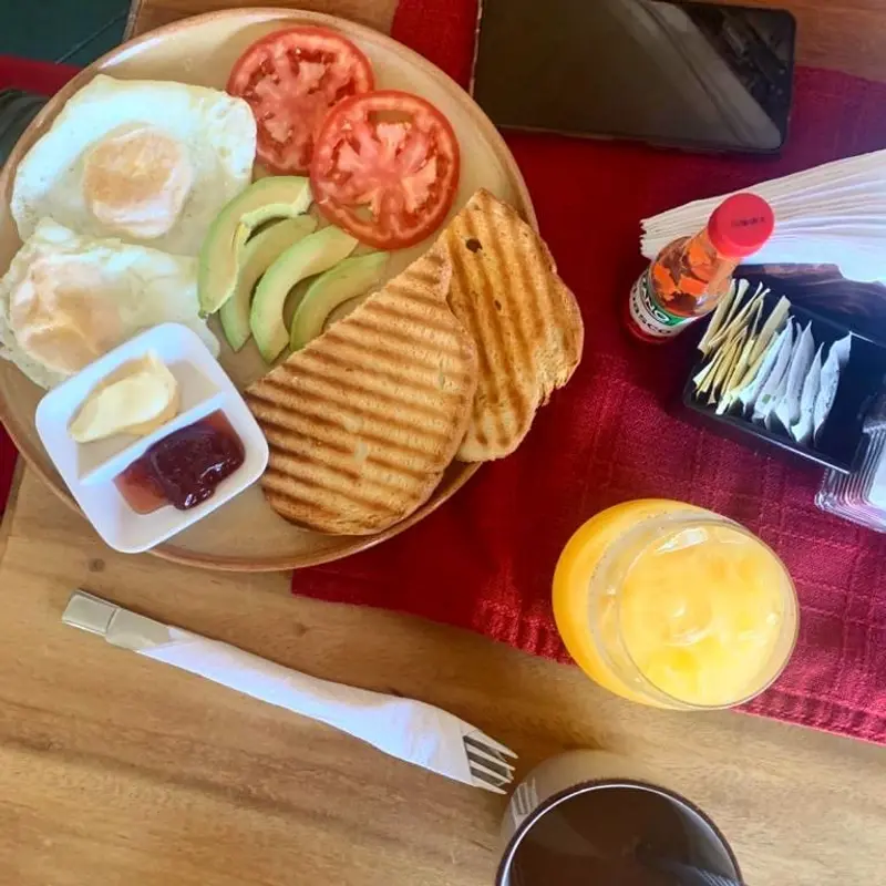 Desayuno con huevos fritos, tomate, aguacate, un sándwich prensado, mermelada y jugo de naranja