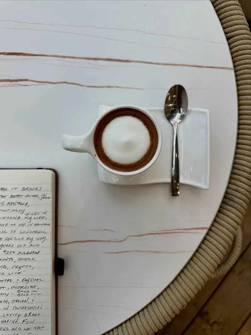 Café pequeño con espuma y cuchara sobre una mesa de mármol, junto a un cuaderno