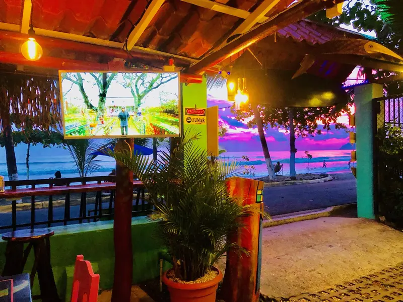 Barra y mesas bajo el techo de palapa al anochecer, con una pantalla y la playa al fondo