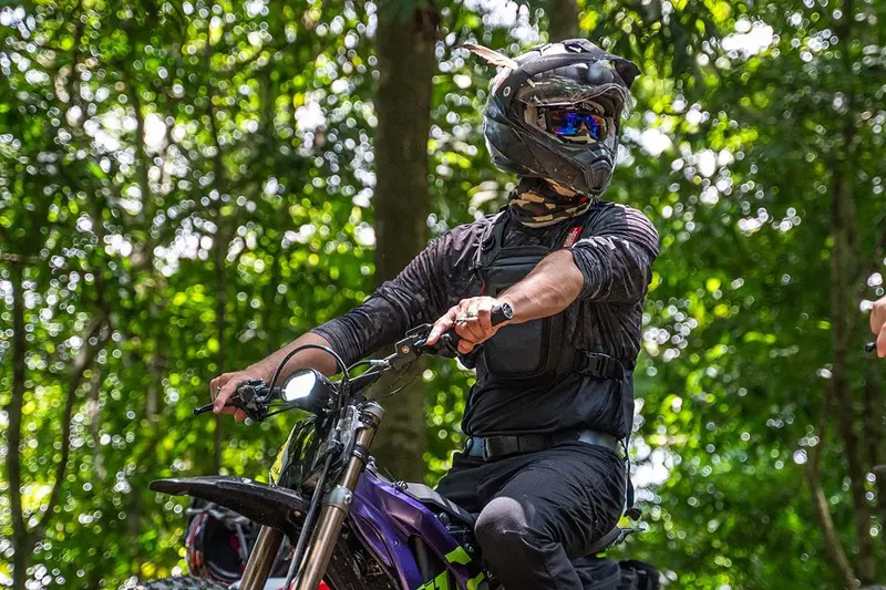 Piloto con casco y chaqueta oscura sobre su moto en un sendero del bosque