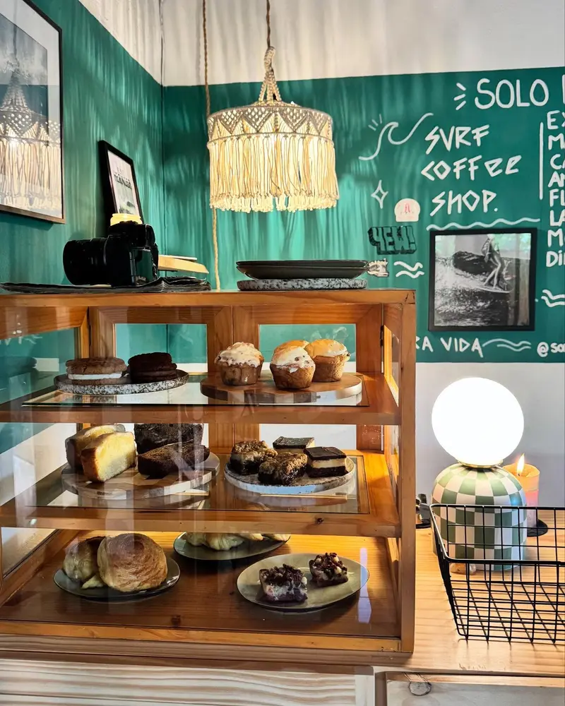 Interior del café con vitrina de repostería, pizarra verde con el nombre del local y lámpara tejida