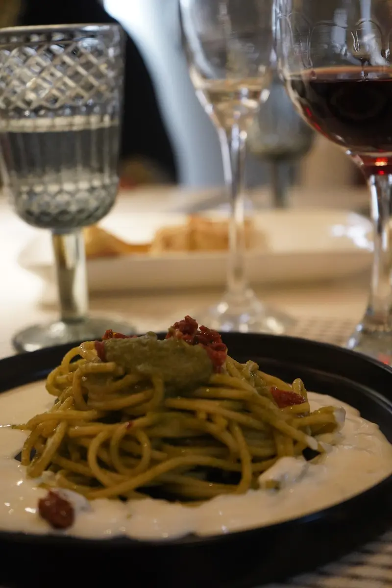 Spaghetti con pesto sobre crema blanca en un plato negro, con copas de vino al fondo