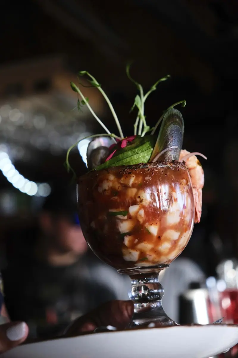 Copa grande de cóctel de mariscos con camarón, mejillones y germinados, sostenida frente a la barra