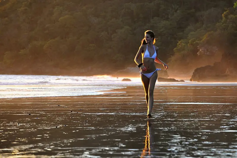 Persona caminando por la orilla al atardecer con luz dorada y rocas al fondo