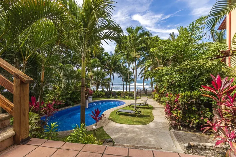 Piscina entre jardines y palmeras con escalera de madera y el mar al fondo
